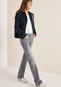 Donna sorridente che cammina, indossando una felpa blu navy con zip, una maglietta bianca, jeans grigi e sneaker bianche, con le mani in tasca, su uno sfondo semplice.
