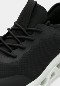 Κοντινό πλάνο μαύρου αθλητικού παπουτσιού με υφασμάτινο επάνω μέρος, μαύρα κορδόνια, ταμπούρα έλξης στη γλώσσα και λευκή σόλα με αεραγωγούς.