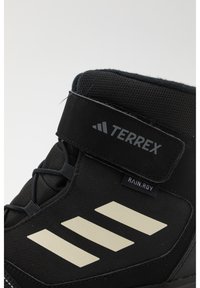 Bota de senderismo negra de caña alta, con parte superior de tejido texturizado, correa ajustable, logo reflectante y tres franjas blancas en el costado.