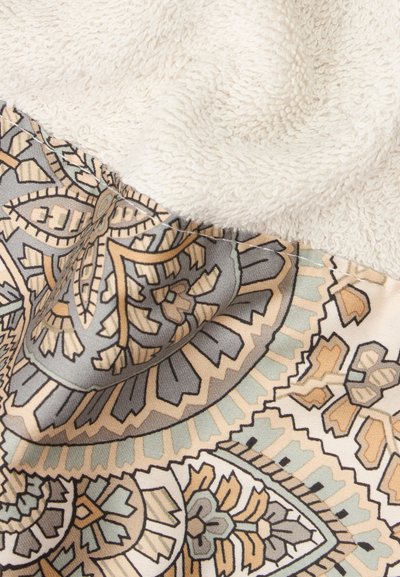 Tissu blanc doux plié sur un textile à motifs avec des formes géométriques et florales beige, grises et bleues.