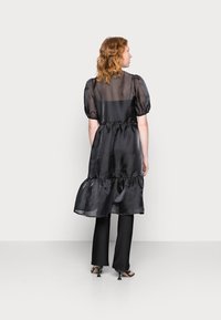 Vestido preto transparente com mangas bufantes, bainha franzida e cintura com franzido. Usado sobre calças pretas ajustadas. Textura suave com um design fluido.