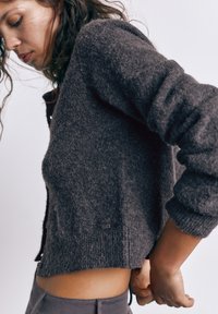 Ung kvinde med krøllet hår iført en mørkegrå uldtrøje og grå bukser, justerer ærmer med hænderne bag ryggen, hvid baggrund.