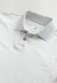 Hvid polo-shirt med en mønstret lysegrå krave, to knapper ved halsen og et mærke med teksten "NEXT", der viser størrelse M. Glat, blød tekstur.