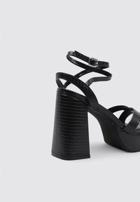 Sandalias de tacón negro con un patrón de cocodrilo texturizado, tacón ancho y correas cruzadas en el tobillo con un detalle de hebilla metálica.