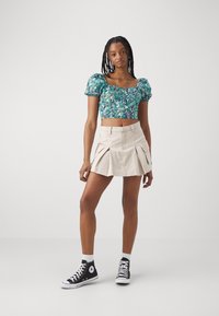 Virágmintás, puffos ujjú zöld és kék tónusú crop top, bézs plisszírozott mini szoknyával és fekete magas szárú tornacipővel párosítva.