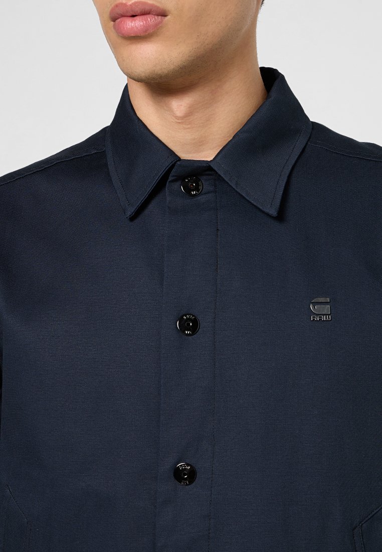 Navy blauwe shirt met een kraag, voorzien van zwarte knopen en een klein logo. Gemaakt van stof met een gladde textuur, ontworpen met een minimalistische esthetiek.