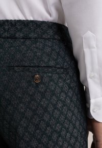 Tmavé texturované kalhoty s knoflíkovou zadní kapsou, které jsou nošeny s bílou košilí s dlouhým rukávem, na které je vidět detail manžet a rukávu.