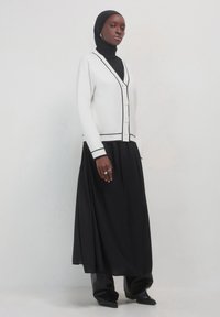 Donna con hijab nero, cardigan bianco con bottoni e rifiniture nere, lunga gonna nera, stivali neri e anelli d'argento sulla mano sinistra.