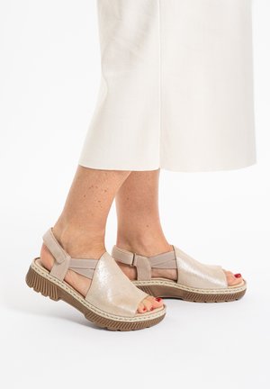 Woman wearing beige open-toe sandals with thick soles and elastic ankle straps, paired with off-white cropped pants.