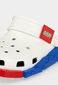 Λευκό παπούτσι τύπου slip-on με οπές αερισμού, ρυθμιζόμενο λουράκι στη φτέρνα και κόκκινη και μπλε σόλα σε στυλ διασυνδεδεμένων τουβλών, με κόκκινη ετικέτα λογότυπου LEGO.