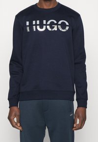 Uomo che indossa una felpa navy con un logo argento lucido "HUGO" sul petto e pantaloni casual blu scuro.