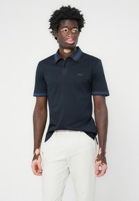 Polo navy com um corpo sólido, colarinho em contraste e detalhes nas mangas, e um logótipo subtil no peito. Combinado com calças claras.