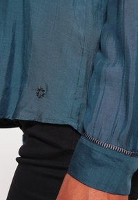 Blusa di seta color teal con una leggera lucentezza, il dettaglio anteriore sinistro mostra un accento decorativo a tre cerchi e un polsino testurizzato con bordi cuciti.