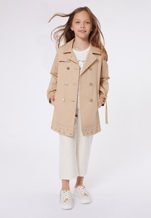 Manteau beige avec des boutons dorés, présentant un design avec des clous décoratifs en bas. Associé à un pantalon blanc court et des baskets.