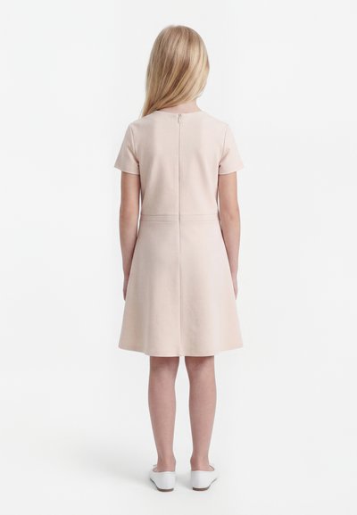 Child with long blonde hair wearing a light beige short-sleeve dress and white shoes, standing facing away against a plain background.