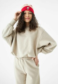 Beige oversized sweatshirt met ribgebreide manchetten, gecombineerd met bijpassende broek. De rode en gestreepte gebreide muts heeft een geborduurde Snoopy-ontwerp.