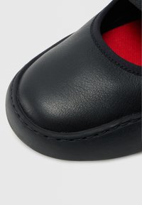 Zapato sintético negro con punta redondeada, superficie texturizada y un interior rojo contrastante. La suela está ligeramente elevada con costuras visibles.