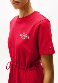 Abito di cotone rosso con vita aderente e maniche corte. Presenta testo del logo bianco e un piccolo accento del logo in blu, rosso e bianco sulla manica.