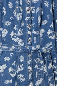 Denimkjole med hvite grafiske trykk av øyne, hender og stjerner. Har knappelukking foran, en knyttet belte og en myk, teksturert overflate.