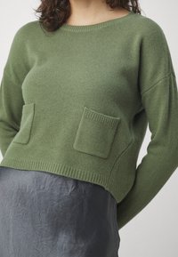 Maglione verde lavorato a maglia con colletto rotondo, due tasche frontali e un design cropped, abbinato a una gonna grigia scura. Texture morbida e bordi a coste.