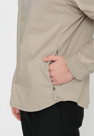 Camicia button-up beige chiaro con texture liscia, tasche laterali e orlo curvo. Presenta bottoni dorati e una cerniera sulla tasca.