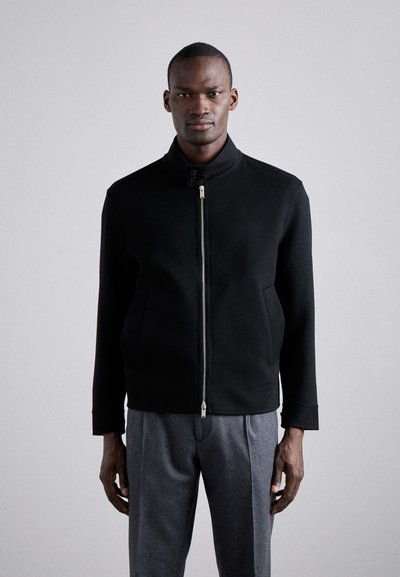 Giacca bomber nera in lana con colletto alto, zip frontale e tasche laterali. Texture liscia e design minimalista. Abbinata a pantaloni grigi.