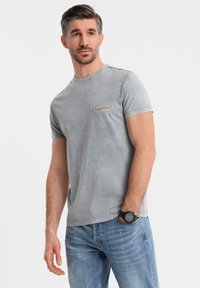 Camiseta gris de manga corta con cuello redondo, que presenta un logotipo en texto "Original" en marrón, combinada con jeans azul claro y un reloj de pulsera negro.
