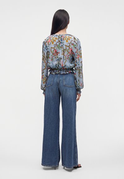 Personne aux longs cheveux foncés de dos, portant un chemisier à fleurs et un jean bleu taille haute à jambes larges, debout devant un fond uni.