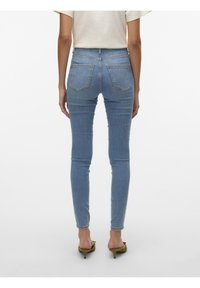 Femme portant un jean skinny bleu clair et des sandales à talons beige, vue de dos sur un fond blanc uni.