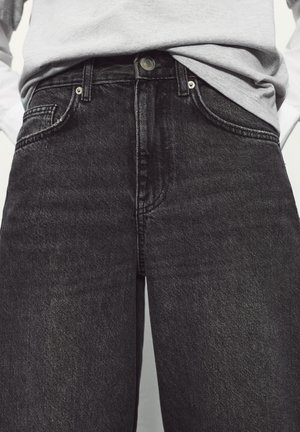 Személy szürke betűrt ingben és magas derekú fekete farmernadrágban, látható gombbal és elülső zsebekkel.