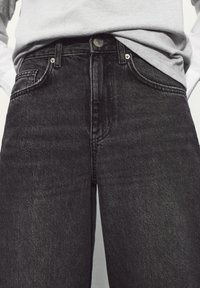 Personne portant une chemise grise rentrée et un jean en denim noir taille haute avec un bouton visible et des poches avant.