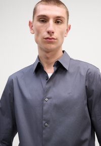 Γκρίζο πουκάμισο με κομψό σχέδιο, δύο κουμπιά, μικρό λογότυπο στο στήθος και λεία υφή. Διαθέτει κολάρο σε σχήμα πιρούνου και πατιλέτα με κουμπιά.