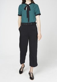 Blusa teal con dettaglio a fiocco e accenti neri, abbinata a pantaloni cropped neri a vita alta e scarpe nere stringate.