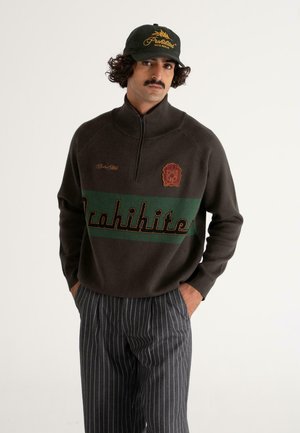 Brauner Reißverschluss-Pullover mit grüner und gelber "Prohibition"-Aufschrift und Logo-Akzenten; kombiniert mit grauen gestreiften Hosen und einer grünen Mütze mit goldener Schrift.