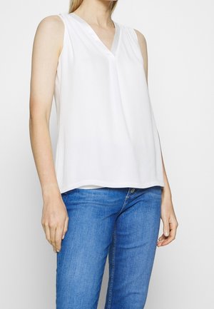 Femme portant un chemisier blanc sans manches à col en V et un jean bleu, debout devant un fond clair uni.