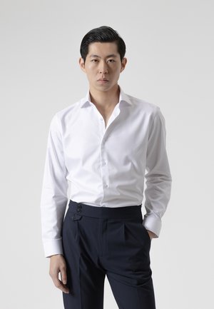 Joven con camisa blanca abotonada y pantalones oscuros está de pie mirando hacia adelante con una mano en el bolsillo sobre un fondo liso.