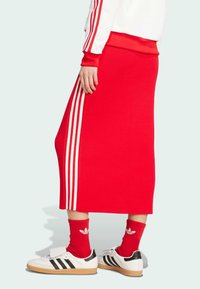 Jupe rouge côtelée avec trois rayures blanches sur le côté, associée à des baskets blanches et noires et des chaussettes rouges, mettant en avant un look sportif.
