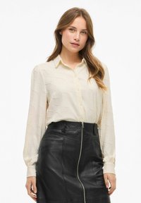 Blusa cremosa e tessurizzata con bottoni frontali e collare, abbinata a una gonna aderente in pelle nera con zip laterale e cuciture dettagliate.