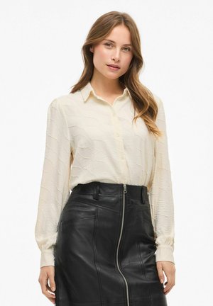 Blusa cremosa e tessurizzata con bottoni frontali e collare, abbinata a una gonna aderente in pelle nera con zip laterale e cuciture dettagliate.