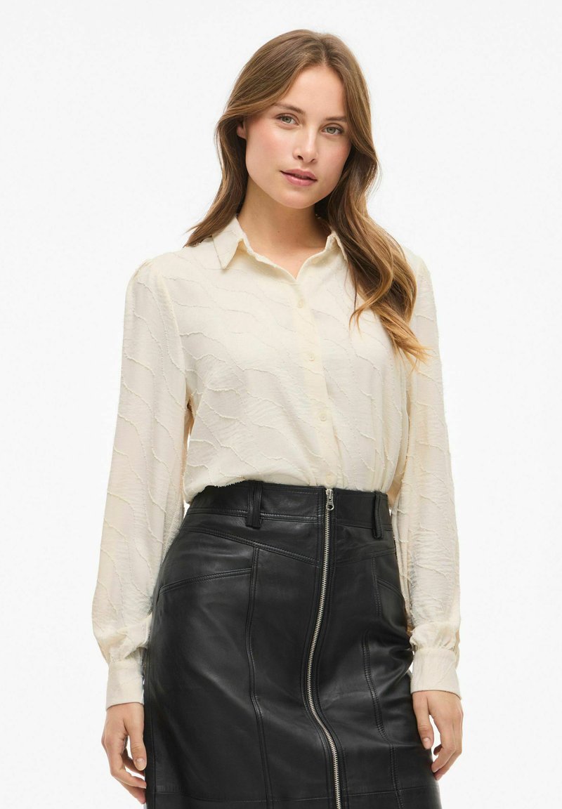 Blusa cremosa e tessurizzata con bottoni frontali e collare, abbinata a una gonna aderente in pelle nera con zip laterale e cuciture dettagliate.