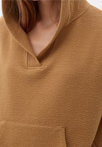 Hnědý svetr s texturovaným povrchem, velkou kapucí, výstřihem do V a přední kapsou. Materiál vypadá měkký a pohodlný.