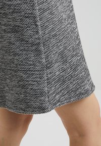 Harmaa ja valkoinen teksturoitu kangashame, jossa on hieman kaareva helma ja geometrinen kuvio. Materiaali vaikuttaa olevan kudottu ja se on keskikokoinen.