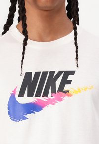 Baltas medvilnės marškinėliai su dideliu juodu "NIKE" logotipu. Po logotipu tęsiasi spalvingas gradientinis swoosh dizainas mėlynos, rožinės ir geltonos spalvų.
