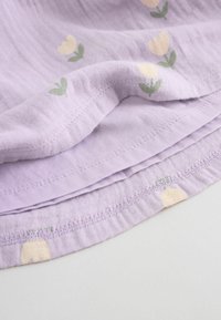 Hellvioletter Stoff mit hellgelben Tulpenmustern und grünen Blättern. Der Rand ist mit einem genähten Saum versehen und hat eine weiche, strukturierte Oberfläche.