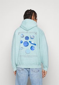 Ljusblå huvtröja med en grafisk design av solsystemet på baksidan, med planeter i olika nyanser av blått och grått.