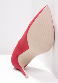 Rode suède hoge hakken met een puntige neus en een slanke naaldhak. Gladde beige buitenzool met textuurlijnen voor grip.