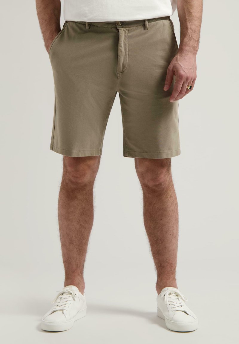Shorts kaki până la genunchi din bumbac, cu față plată, bucle pentru curea, două buzunare laterale și o textură netedă. Purtați cu adidași albi.