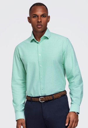 Jeune homme aux cheveux courts portant une chemise boutonnée vert menthe, une ceinture tressée marron et un pantalon bleu foncé, debout devant un fond uni.