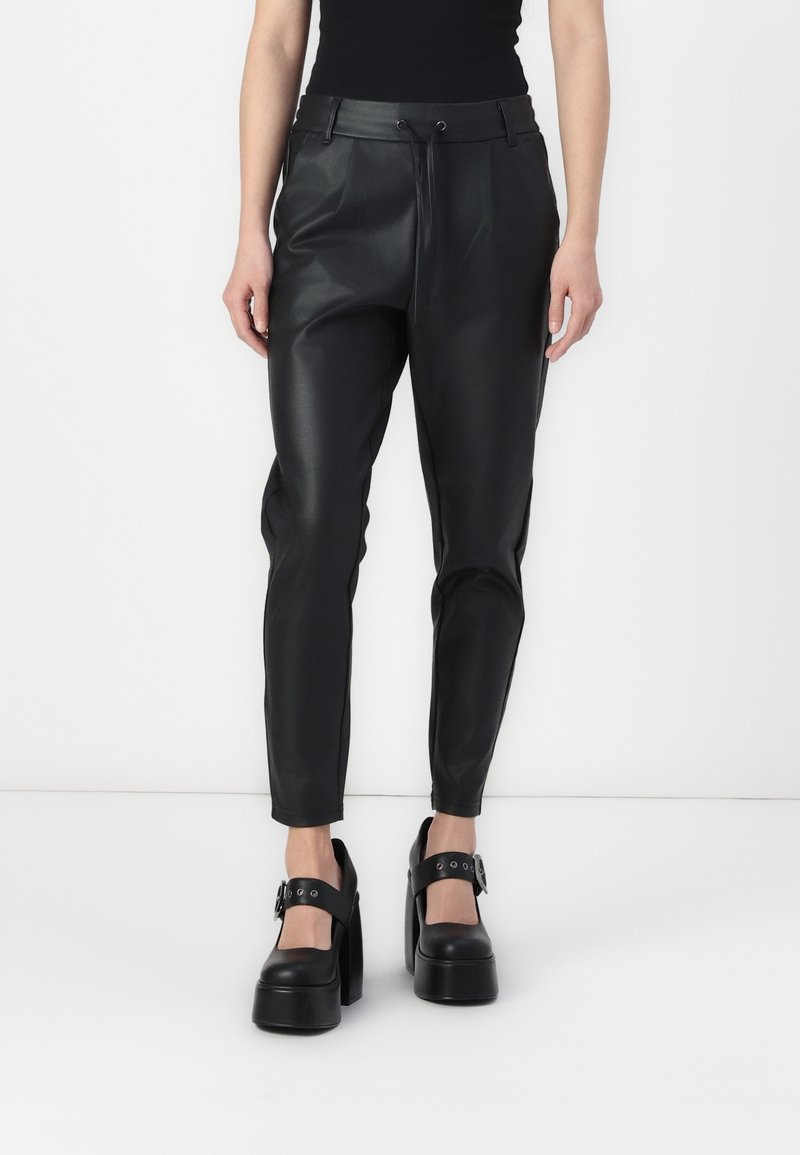 Femme portant un pantalon en cuir noir ajusté avec une taille à cordon de serrage et des chaussures à plateforme à talons hauts noires avec des brides à boucle, debout devant un fond blanc.