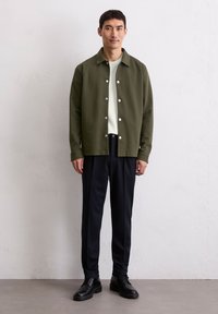 Oliwkowa kurtka zapinana na guziki na jasnoszarym podkoszulku, w połączeniu z ciemnoniebieskimi spodniami i czarnymi sznurowanymi butami. Gładka faktura materiału.